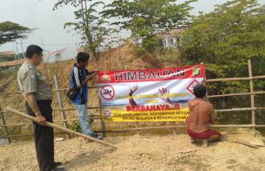 Bhabinkamtibmas Desa Wibawa Mulya Aiptu Nasichin dibantu dengan anggota Karang Taruna Desa Wibawa Mulya saat memasang spanduk himbauan dilarang berenang dan bermain air di kubangan yang berada di dekat Perumahan KSB tepatnya di Kp. Tegal Panjang RT 01/01, Desa Wibawa Mulya, Kecamatan Cibarusah.